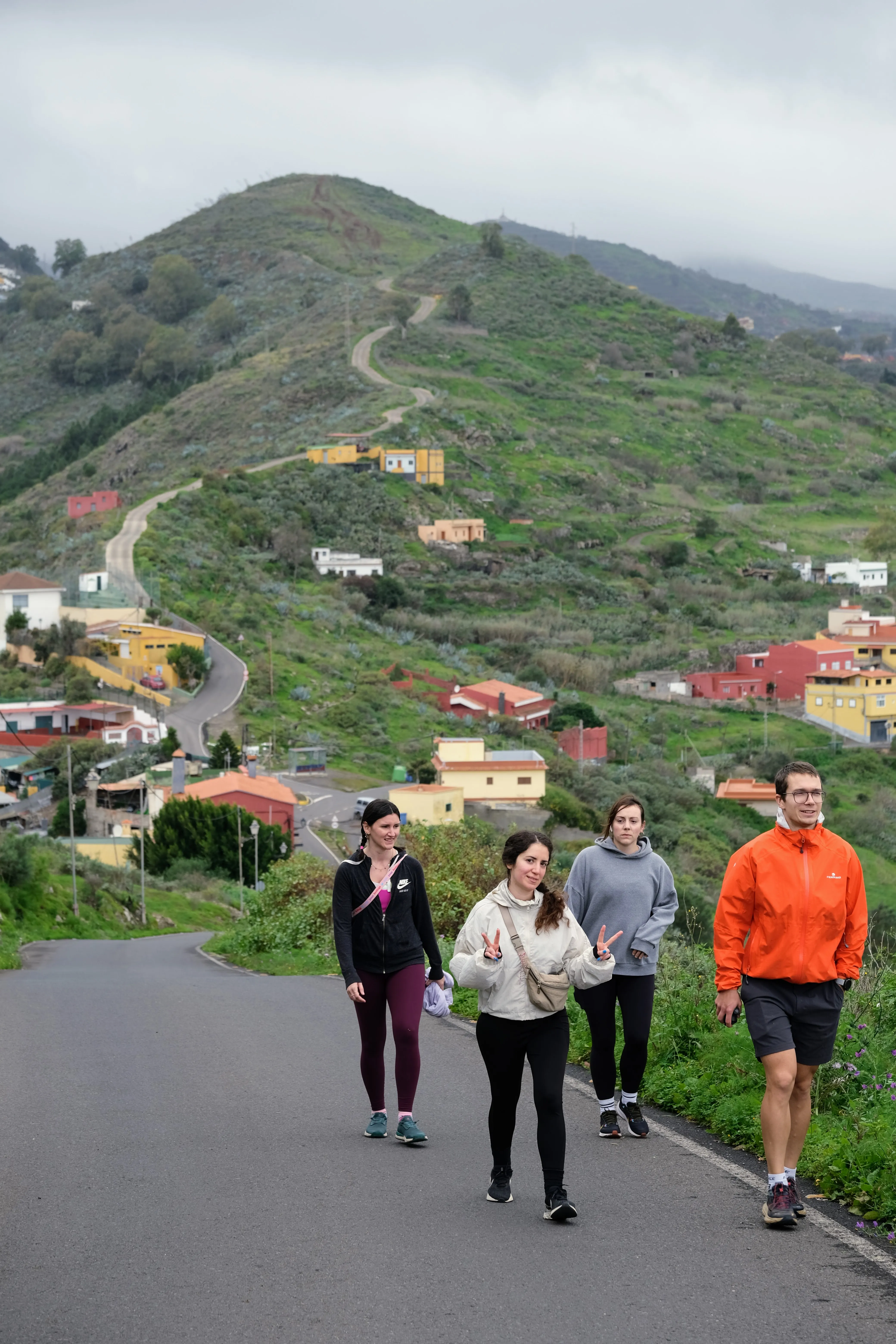 Jillian, Iris, Ena and Mat walk towards the camera on a paved road. In the background, the road winds up a hill covered in lush vegetation and colorful houses dotted throughout.