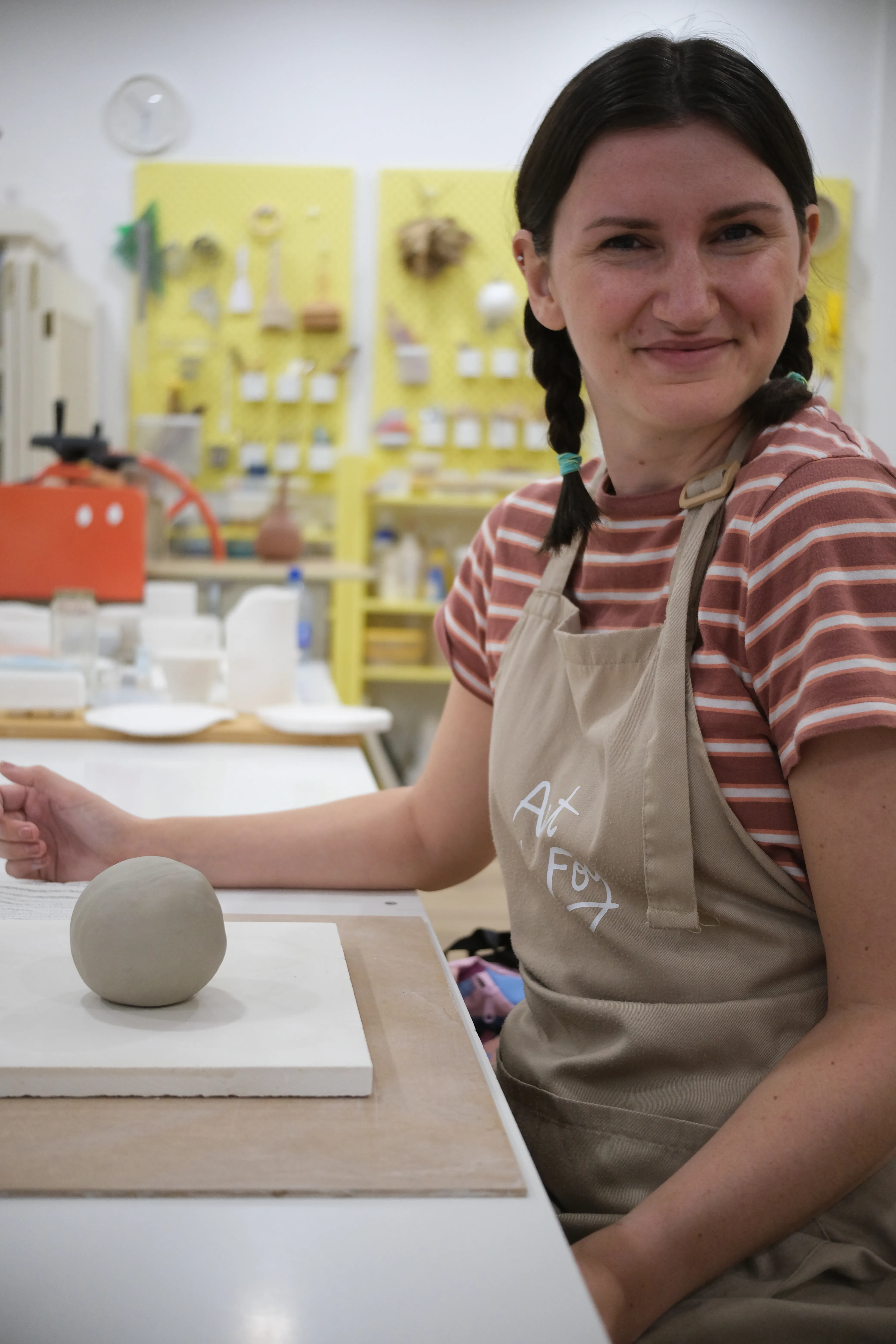 Closeup of Jillian sitting in front of a small ball of clay. She is wearing an ocher apron and a cute brown-and-white striped t-shirt.