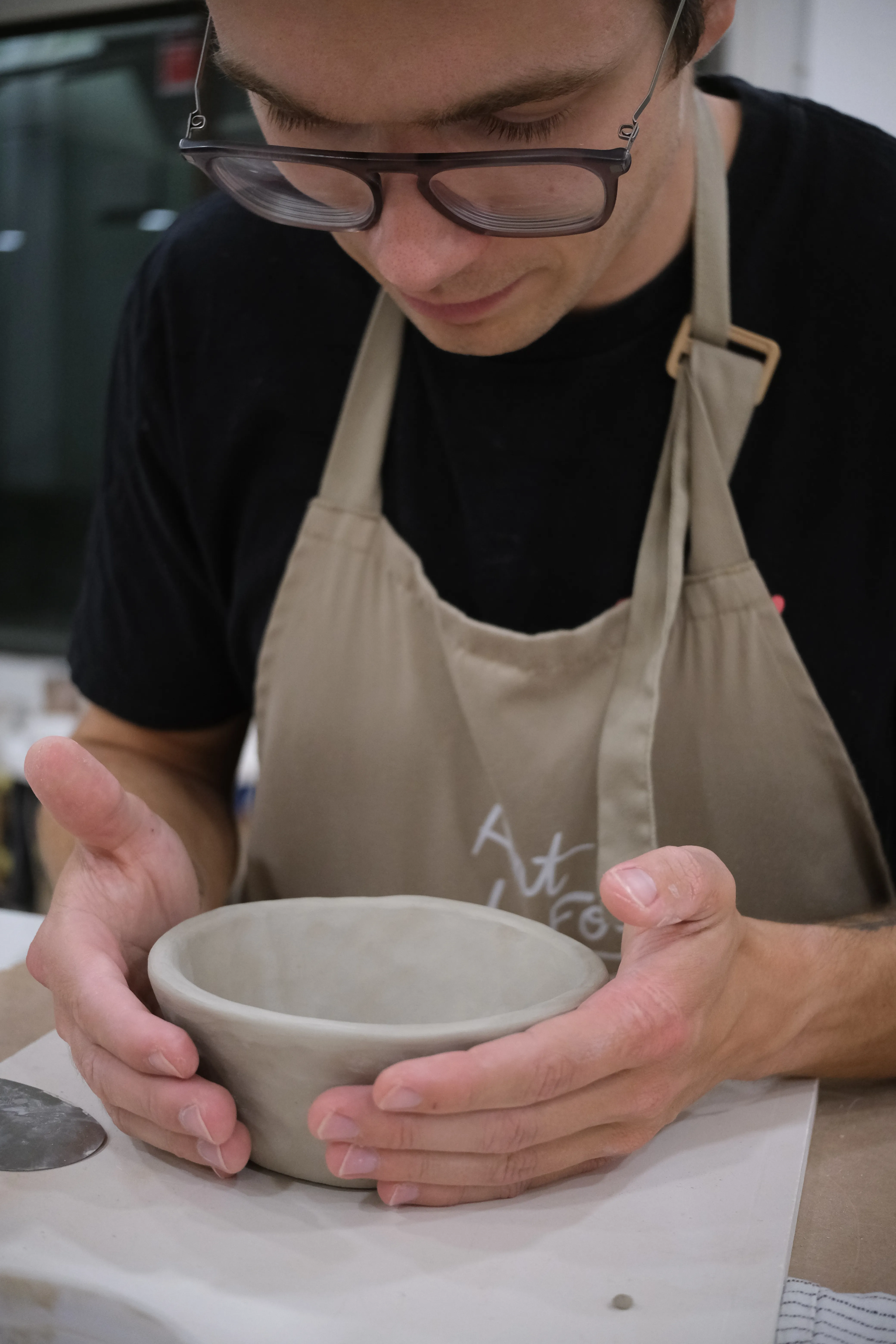 Closeup of Mat gently cradling a bowl of clay in his hands. He is wearing black-rimmed glasses, a black t-shirt and an ocher apron.