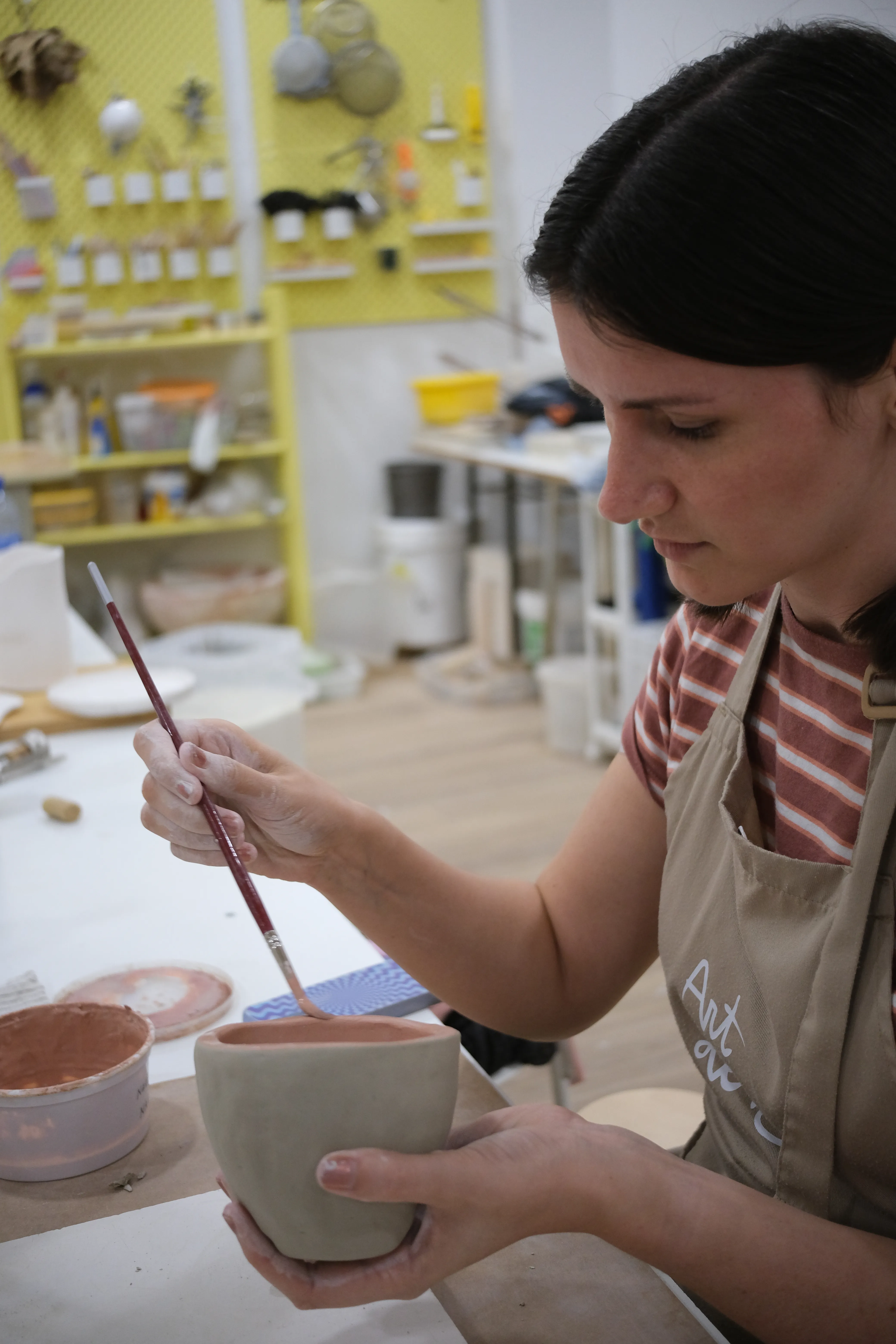 Jillian holds a mug of clay in her left hand and carefully applies a coat of pastel pink paint with a long, thin brush in her right hand.