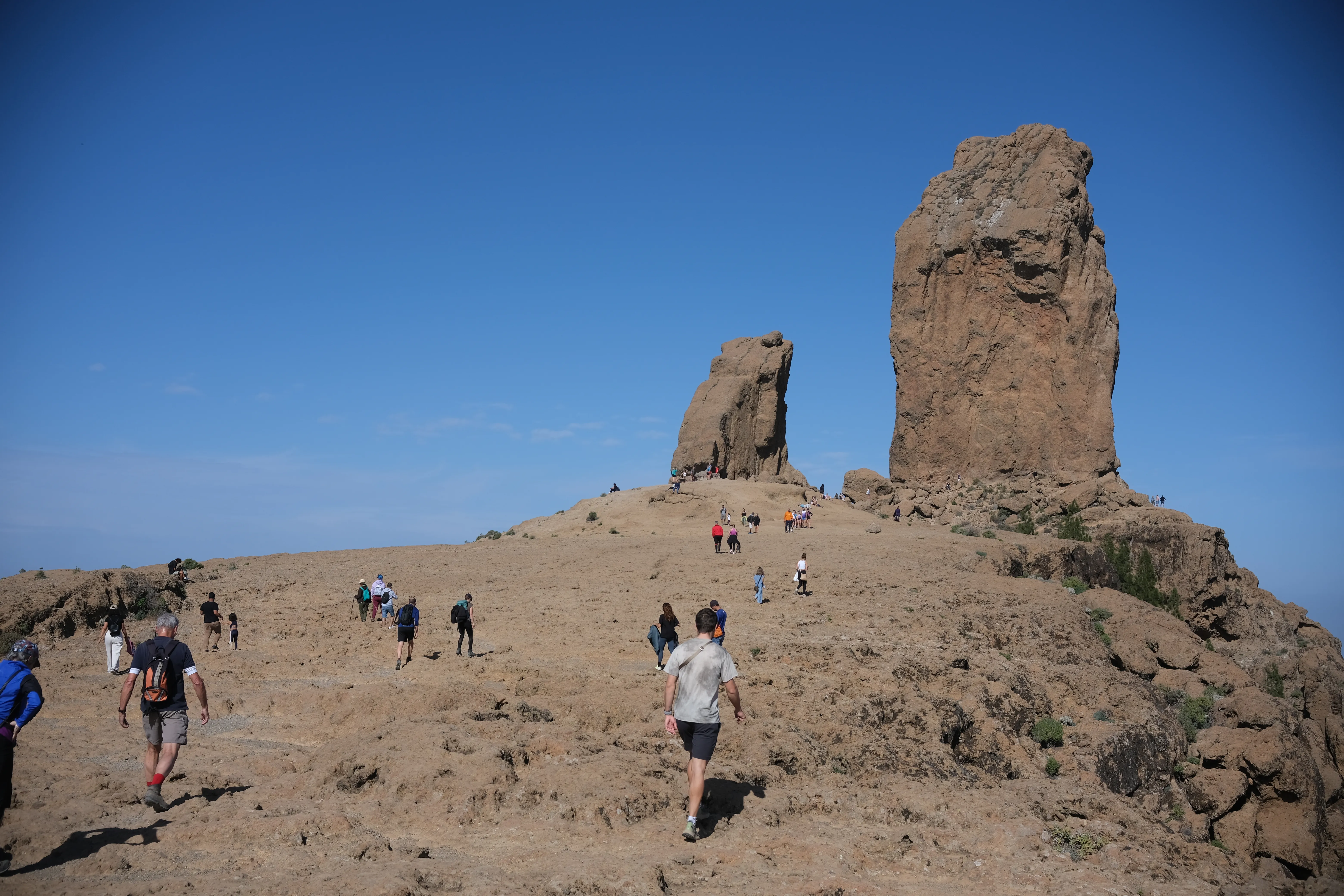 Wide shot of a flat, barren moutaintop. Groups of hikers walk towards two imposing rock columns jutting into the blue sky.
