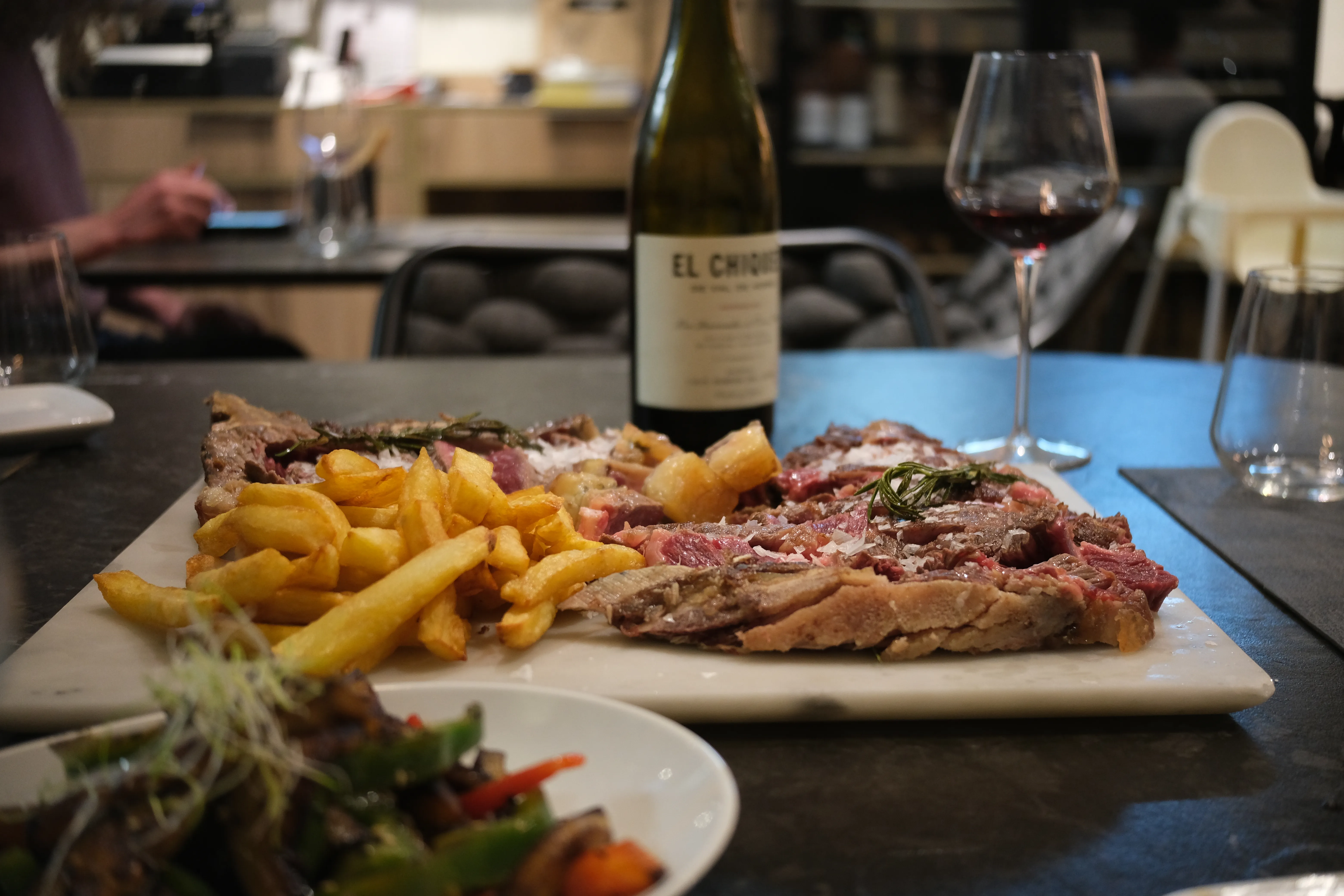 A fancy table at a restaurant with a plate of sliced, pink dry-aged beef with flaked salt and fries in the center, a bowl of salad in the foreground and a bottle of red wine in the background.