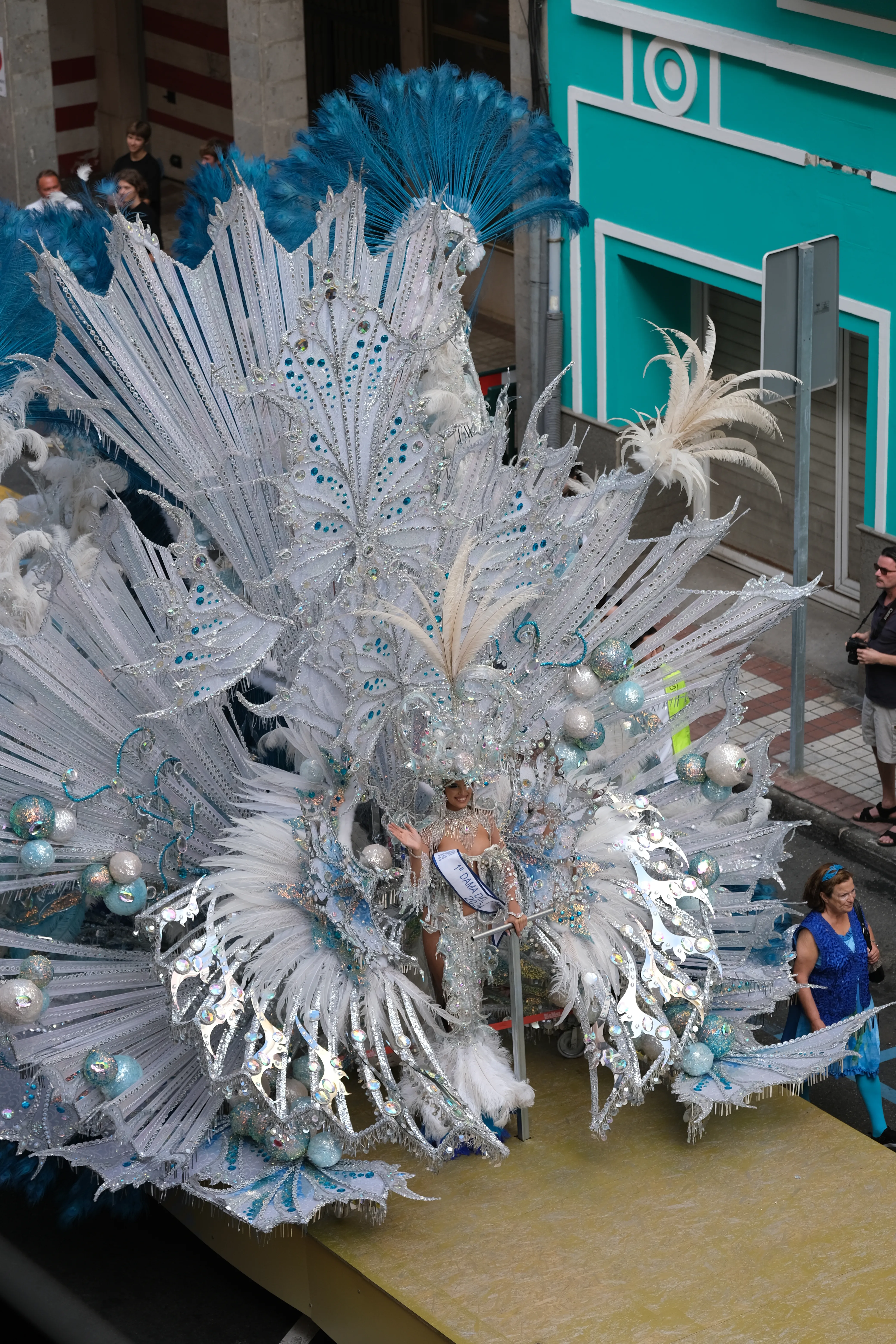 A woman stands atop a carnival float wearing an extravagant and enormous silver costume shaped like pecock feathers.