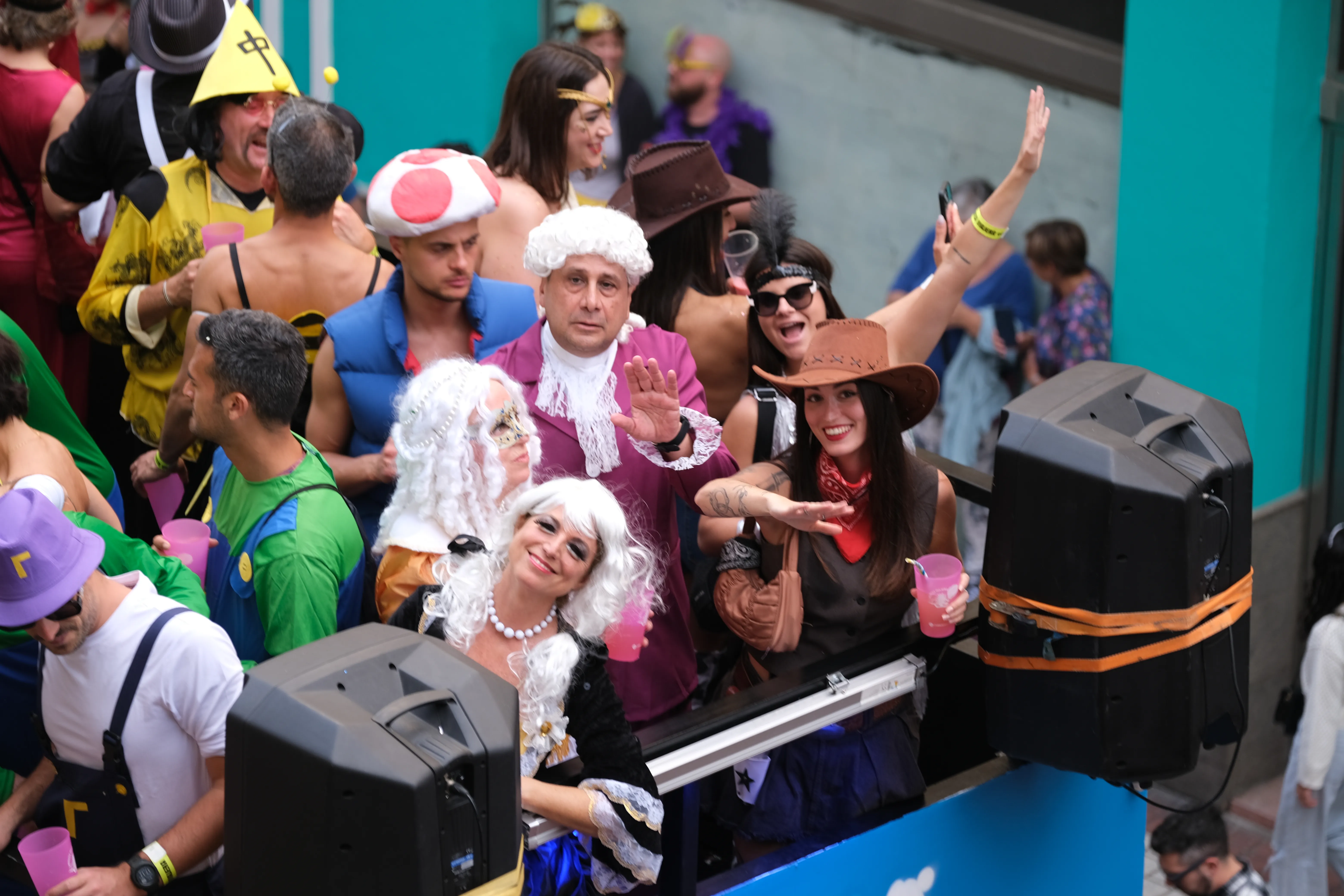 A group of people dressed up in various carnival costumes stand tightly packed atop a party bus and wave at the camera.