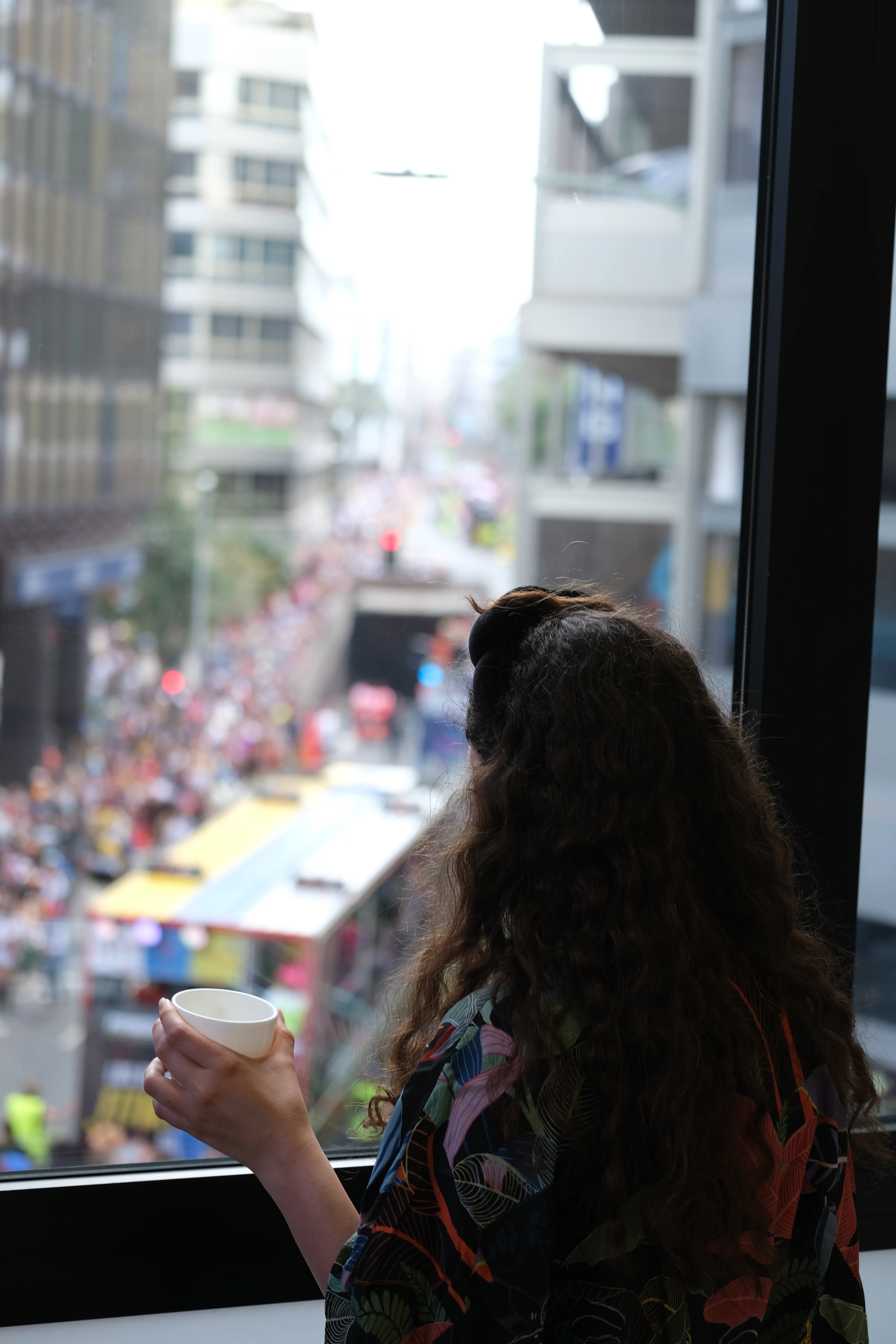 Iris stands in front of a window holding a cup and watches the carnical spectacle on the street below.