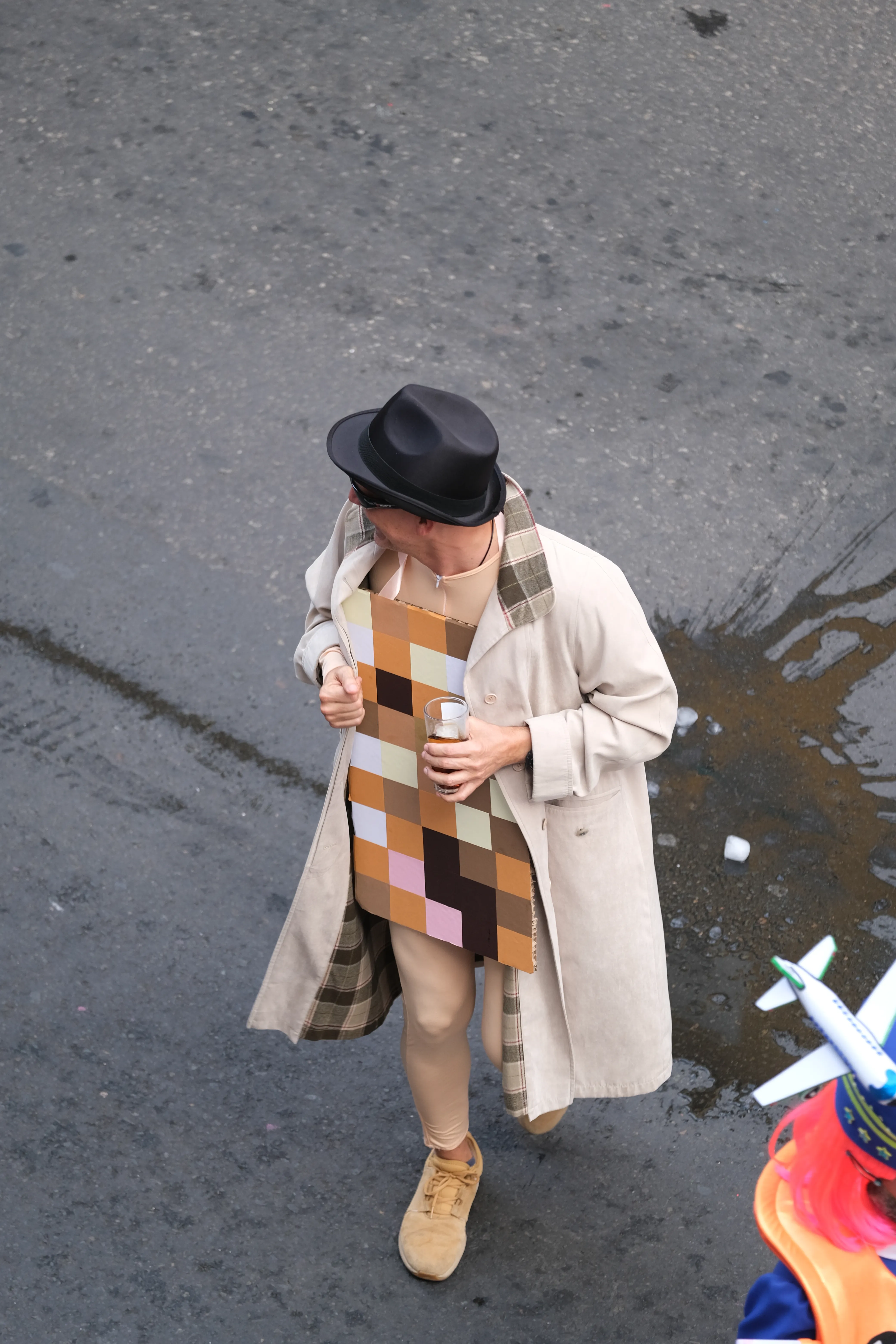 A man wears a beige coat that is open in the front to reveal a piece of cardboard styled to look like a pixelated, censored image of his bare body.