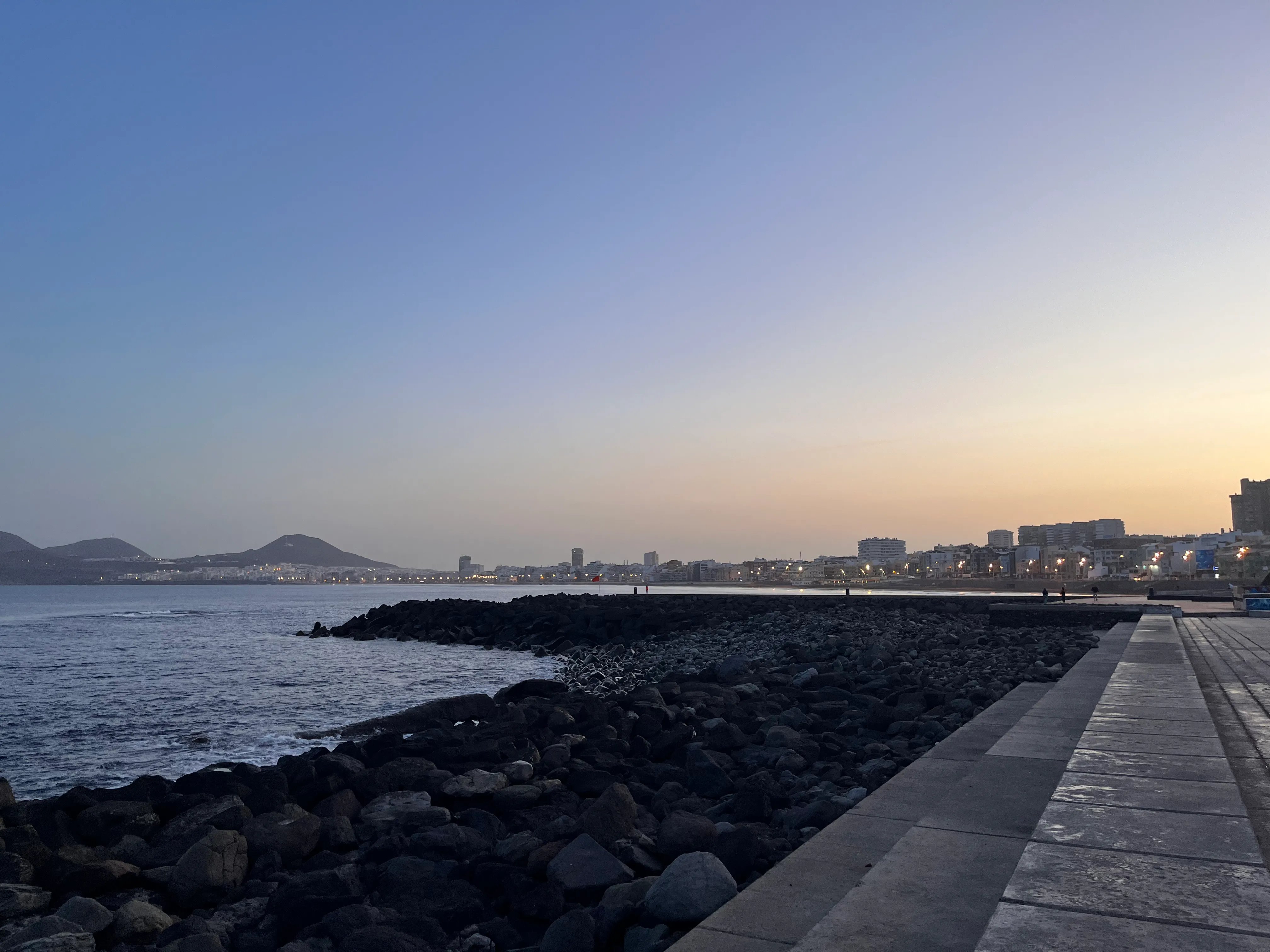 A rocky shoreline reaches out into the dark blue ocean waters in the grey-blue morning twilight. In the background, lights awaken in the city of Las Palmas.